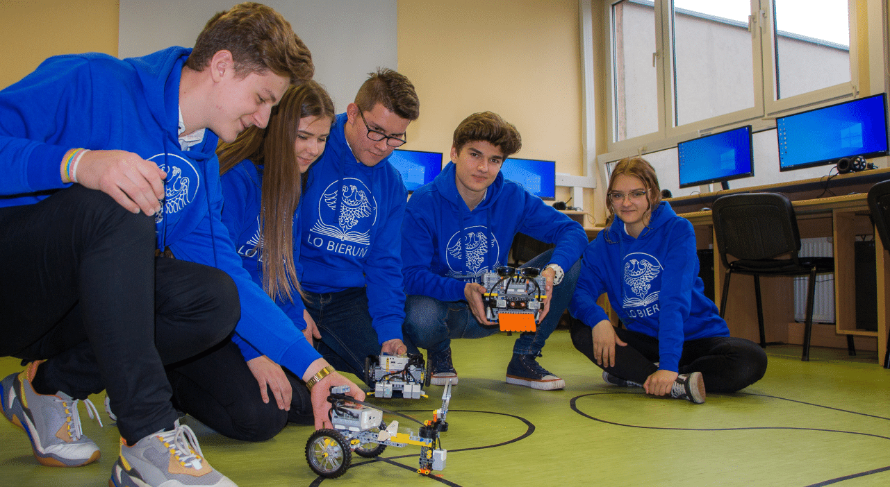 Na zdjęciu uczniowie Liceum w Bieruniu ubrani w bluzy z logo szkoły kucają trzymając w rękach roboty zaprogramowane do poruszania się po określonej trasie.